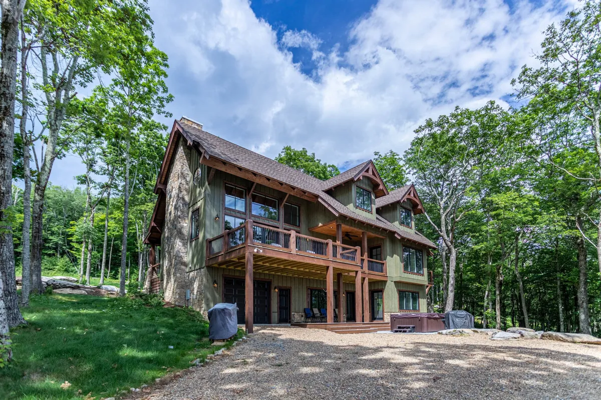 A welcoming exterior view of the house showcases its gabled roof, expansive deck, and mature trees surrounding the structure. The scene exudes a sense of privacy, spaciousness, and harmony with nature.