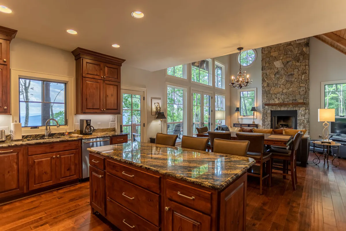 An open-plan area featuring a large kitchen island, a stone fireplace, big windows with views, and a dining space seamlessly connected to the living room. The arrangement encourages social interaction and shared experiences.