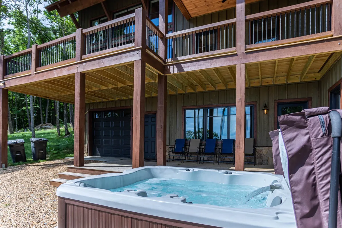 The spacious wooden deck features a hot tub overlooking a serene natural landscape, with large windows connecting the indoor and outdoor living areas. The inviting setup suggests relaxation after a day of outdoor activities.