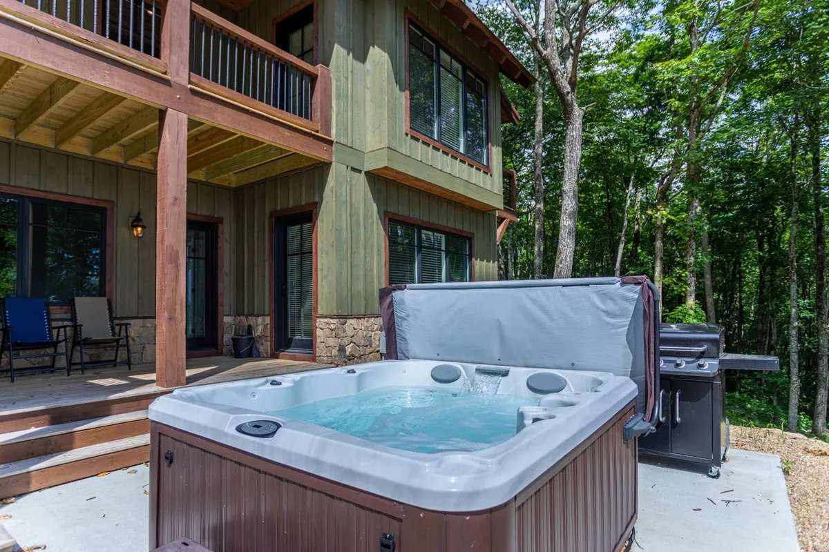 A spacious wooden deck nestled among trees, equipped with a hot tub and grill. The setting is perfect for families or groups relaxing outdoors, socializing, or enjoying a barbecue amid tranquil, natural surroundings.