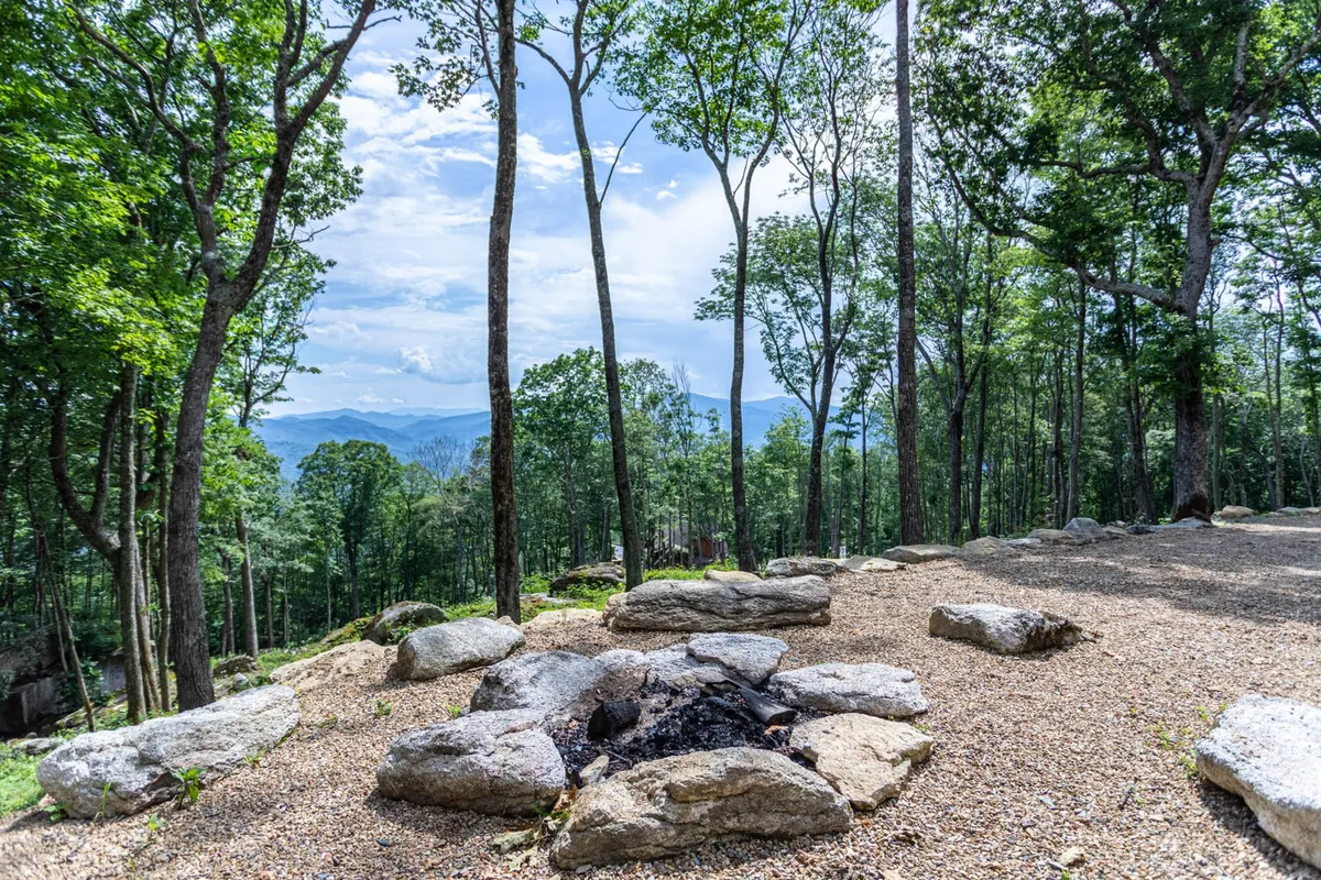 A rustic fire pit is encircled by trees with a mountain vista in the distance, creating a cozy spot for gatherings, roasting marshmallows, and stargazing. The ambiance is relaxed and invites guests to linger outside.