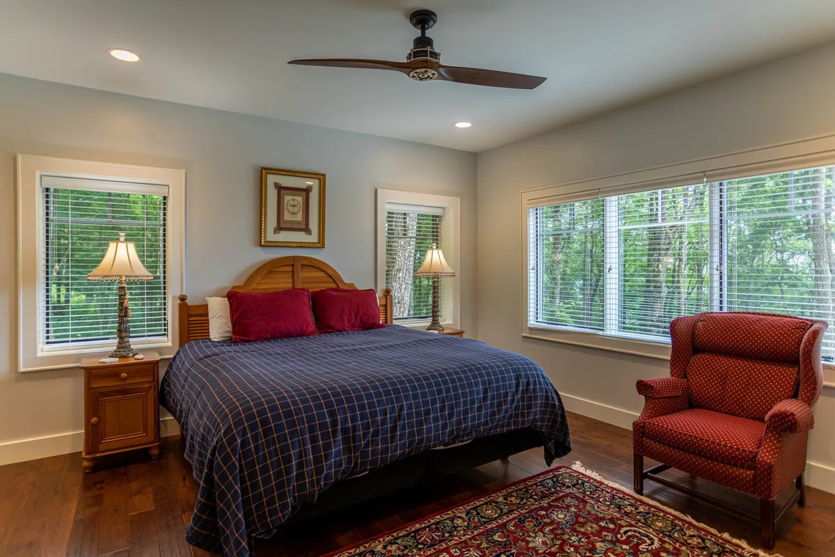 A cozy guest bedroom with blue checkered bedding, red accent pillows, and a wooden nightstand with a lamp. A red upholstered armchair is positioned near large windows, filling the room with natural light and offering a peaceful view. The space is welcoming and perfect for relaxation or family stays.
