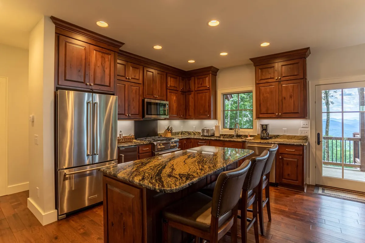 A warm, inviting kitchen featuring a prominent granite countertop at the island, wood cabinetry, ample island seating, and a large window that provides both natural light and scenic views. The space is clearly designed for social cooking and gathering, with open sight lines that encourage group participation.