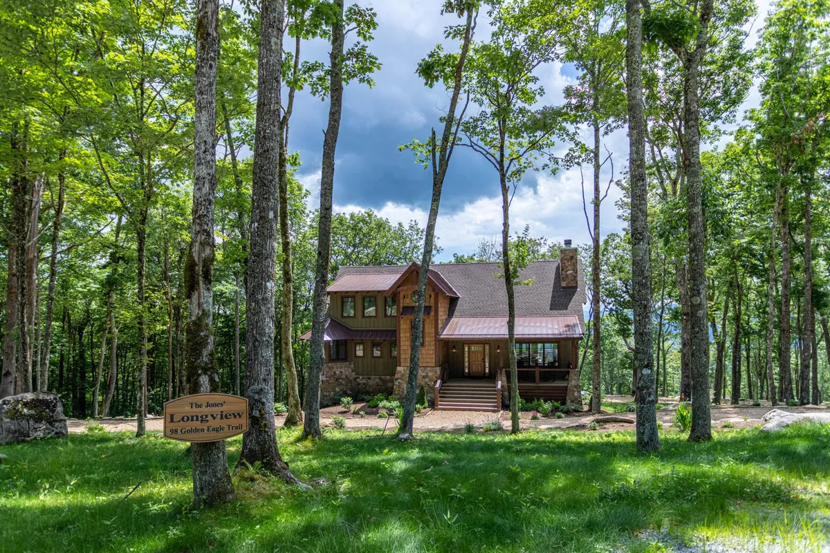 A wooden house sits nestled among trees, with a signpost near a grassy area, inviting guests to explore the peaceful surroundings. The scene suggests easy access to trails and a welcoming environment for group activities and dog walks.