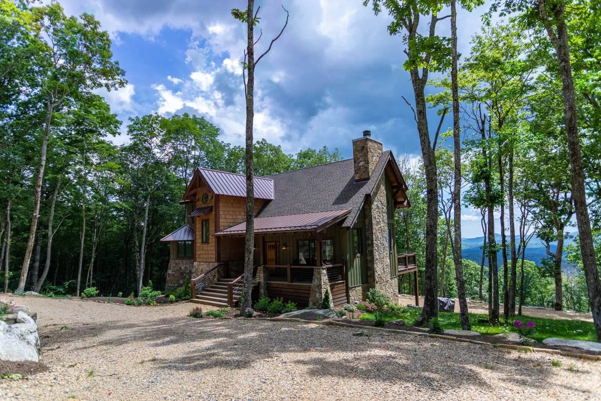 This exterior image captures the cabin's wooden structure with a stone facade, large windows, and is surrounded by trees and a gravel driveway. The setting suggests a serene mountain atmosphere that beautifully transitions through the seasons.