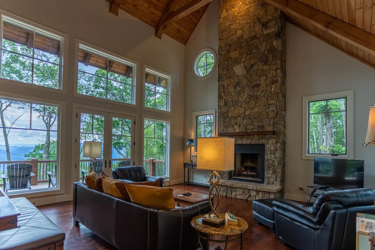 A spacious living room with a stone fireplace as the focal point, large windows that frame scenic mountain views, and an inviting seating arrangement. The natural light streaming in and comfortable furniture create a warm, welcoming setting.