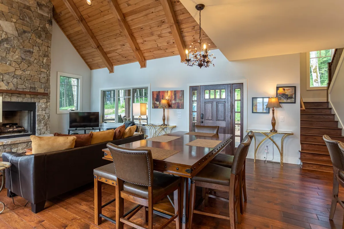 A welcoming dining area featuring a sturdy wooden dining table, large windows offering plenty of natural light, a stone fireplace that adds ambiance, and exposed wooden beams overhead. The space feels both intimate and perfect for group feasts or celebrations.