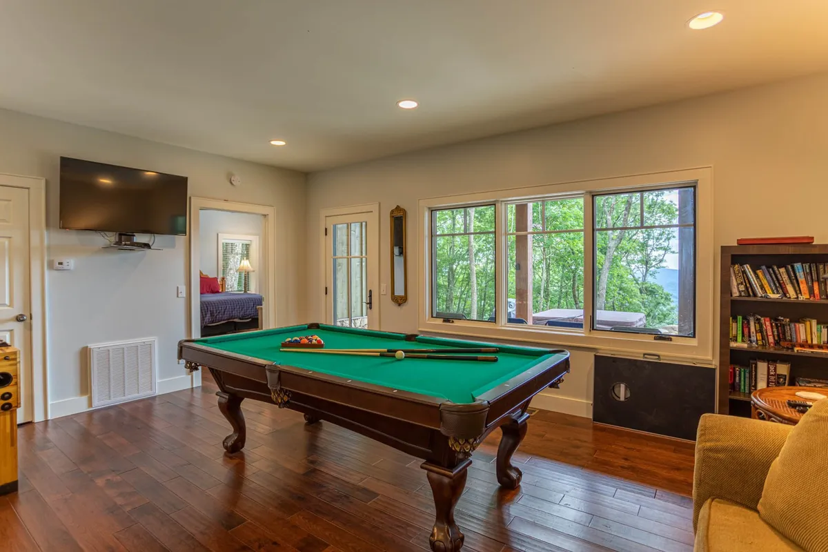 The image centers on a pool table with a backdrop of a large window offering beautiful scenic views and a bookshelf nearby. The well-lit room radiates friendliness and recreation, inviting guests to enjoy a relaxed game or unwind while admiring the view.